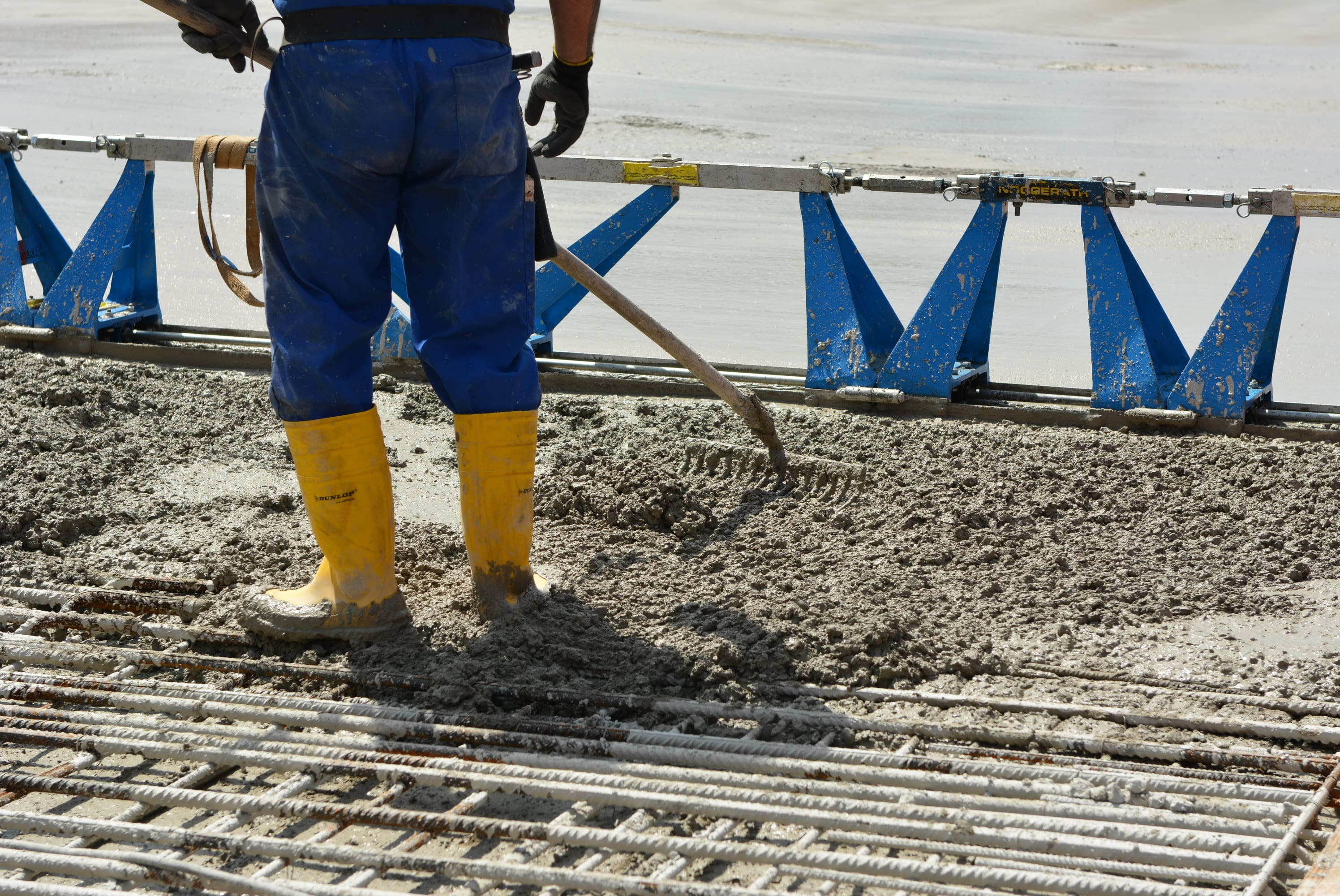 Ein Arbeiter in blauer Hose und gelben Gummistiefeln verteilt Beton mit einer Schaufel auf einer Baustelle. Im Vordergrund ist ein Gitter aus Stahlstäben, im Hintergrund blaue Metallstützen. Der Beton ist noch nass.
